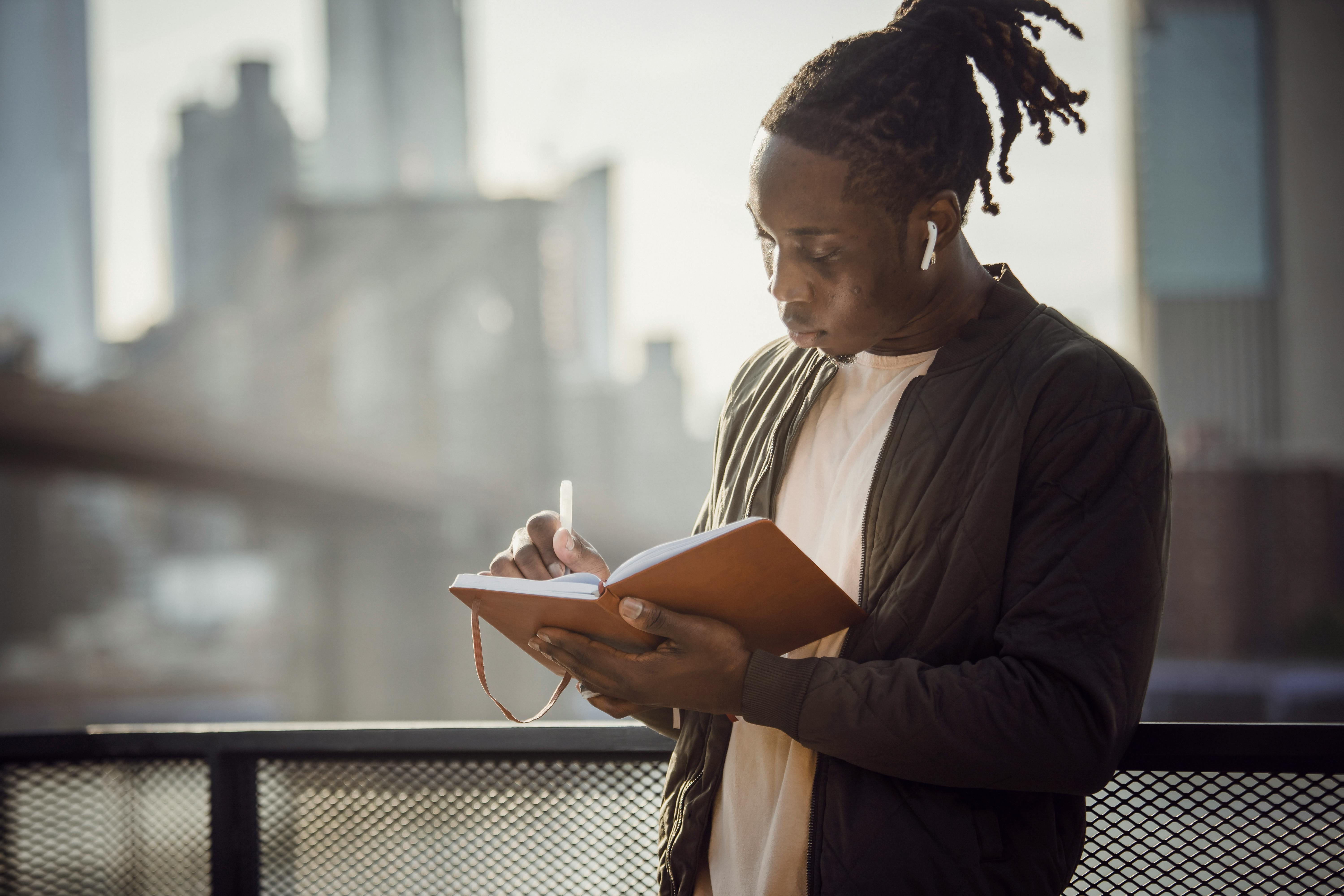 A young man writing in a notebook outdoors with city skyline in the background.
